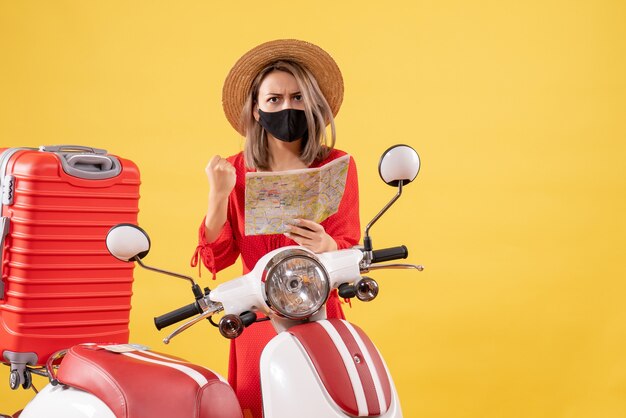 Vista frontal de uma jovem zangada com uma máscara preta segurando um mapa perto de uma motocicleta