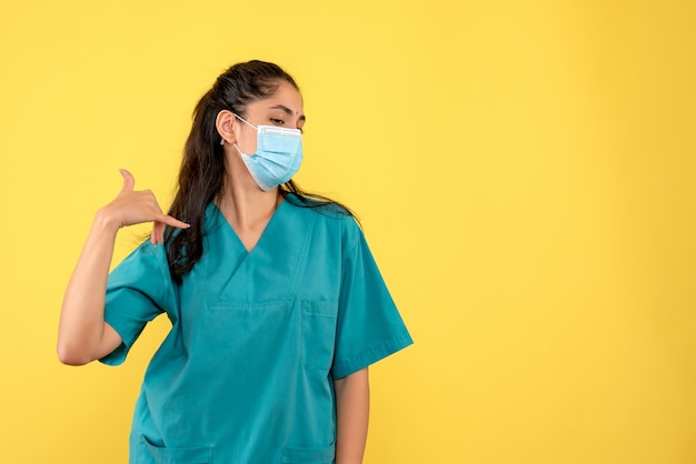 Vista frontal de uma médica bonita com máscara médica, fazendo um gesto de telefonema na parede amarela