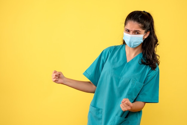 Vista frontal de uma médica bonita com máscara médica mostrando gesto vencedor na parede amarela