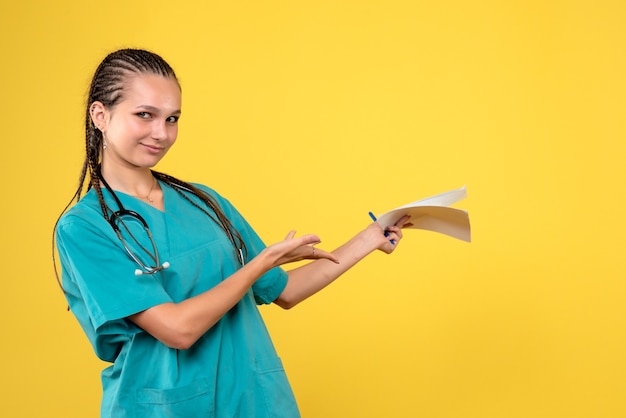Vista frontal de uma médica em um terno médico com papéis na parede amarela