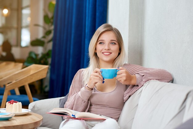 Vista frontal de uma menina loira sorridente, bebendo um café saboroso, sentada no sofá branco