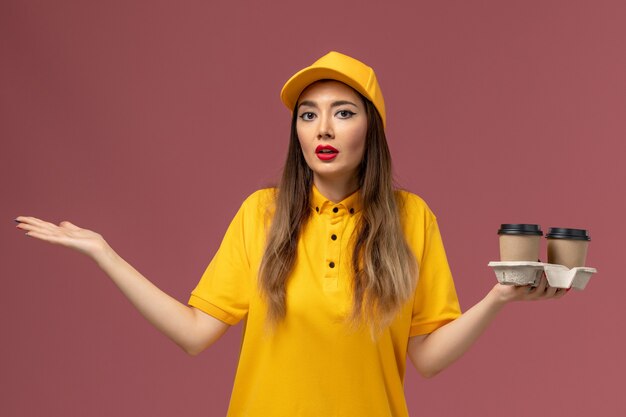 Vista frontal de uma mensageira de uniforme amarelo e boné segurando xícaras de café na parede rosa