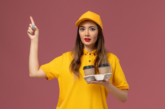 Vista frontal de uma mensageira de uniforme amarelo e boné segurando xícaras de café na parede rosa