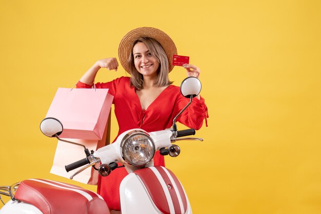 Vista frontal de uma mulher alegre em um vestido vermelho na motocicleta segurando sacolas de compras e um cartão