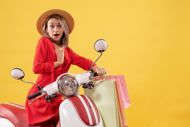 Vista frontal de uma mulher loira com um vestido vermelho em uma motocicleta segurando sacolas de compras e apontando para ela mesma
