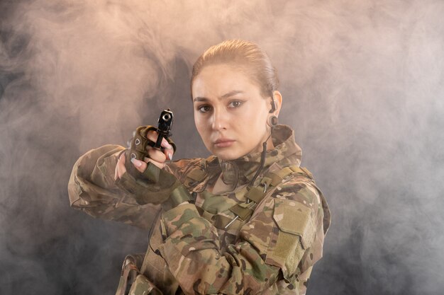 Vista frontal de uma mulher soldado com arma de uniforme na parede preta esfumaçada