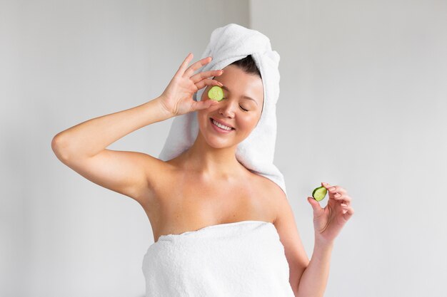 Vista frontal de uma mulher sorridente com uma toalha na cabeça segurando fatias de pepino