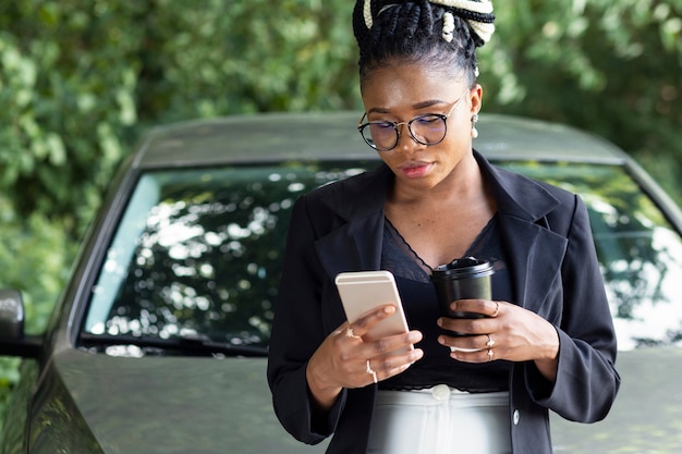 Foto grátis vista frontal de uma mulher tomando café e olhando para o smartphone enquanto se encosta em seu carro