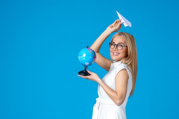 Foto grátis vista frontal de uma trabalhadora vestida de branco segurando um globo terrestre e um avião de papel na parede azul