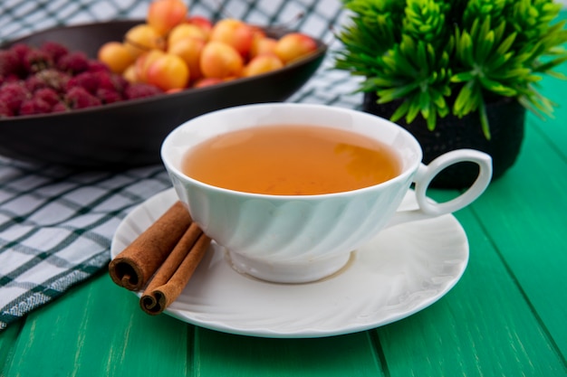 Vista frontal de uma xícara de chá com framboesas com canela e cerejas brancas em uma superfície verde