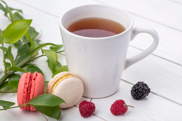 Vista frontal de uma xícara de chá com framboesas e macarons com galhos de folhas em uma superfície branca