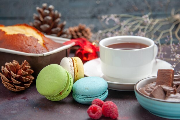Vista frontal deliciosos macarons franceses com chocolate e uma xícara de chá no fundo escuro, chá, bebida, torta, biscoito, bolo, biscoitos