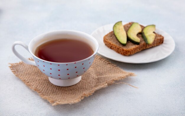 Vista frontal do abacate fatiado saudável em uma lâmina de pão em um prato branco com uma xícara de chá no pano de saco na superfície branca
