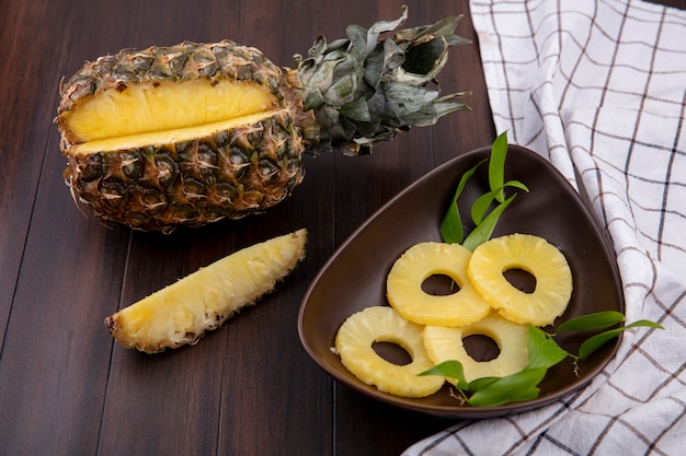 Vista frontal do abacaxi com um pedaço cortado de frutas inteiras e tigela de fatias de abacaxi no pano xadrez e superfície de madeira