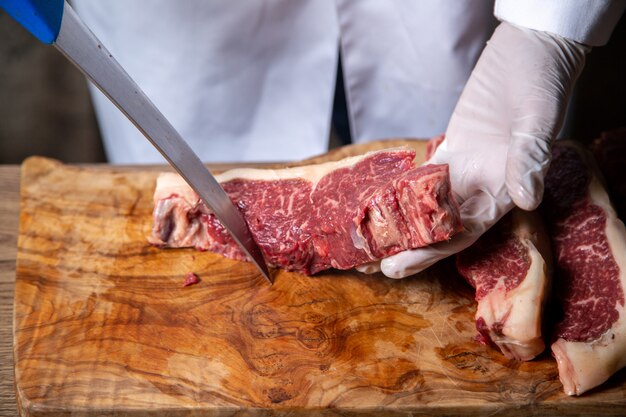 Vista frontal do açougueiro cortar carne em luvas brancas, segurando uma faca grande na mesa de madeira