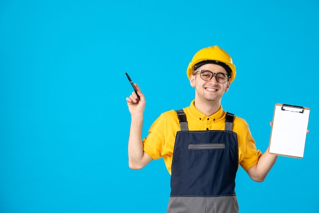 Vista frontal do alegre construtor masculino de uniforme com uma nota de arquivo nas mãos na parede azul