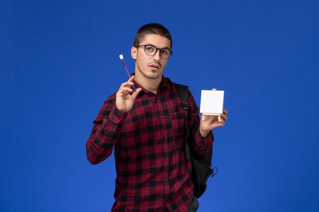 Vista frontal do aluno do sexo masculino com camisa quadriculada vermelha com mochila segurando a borla e cavalete na parede azul