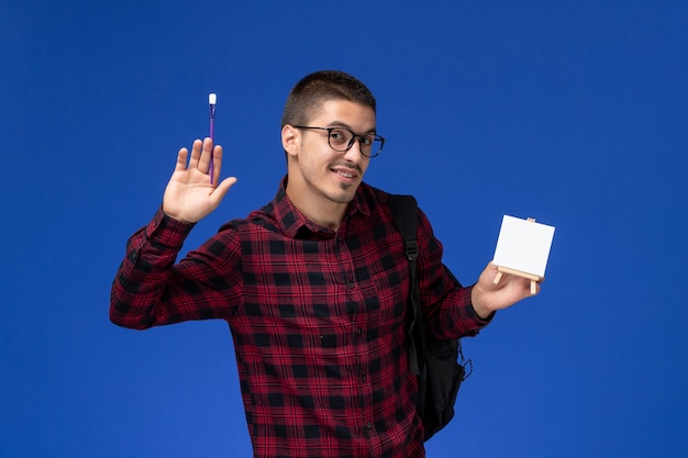 Vista frontal do aluno do sexo masculino com camisa quadriculada vermelha com mochila segurando a borla e cavalete na parede azul