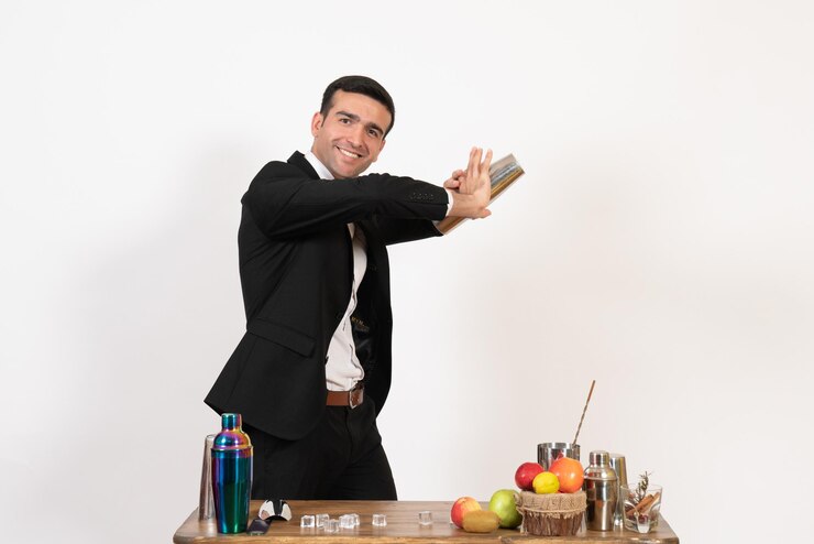 Foto grátis vista frontal do barman masculino de terno em pé em frente à mesa com coquetéis e fazendo uma bebida na parede branca do clube noturno masculino.