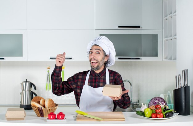 Vista frontal do chef masculino apontando para trás em pé atrás da mesa da cozinha na cozinha