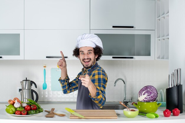 Vista frontal do chef masculino feliz e positivo com legumes frescos e cozinhando com utensílios de cozinha e apontando para a frente e para cima na cozinha branca