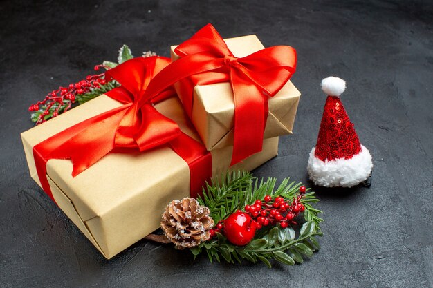 Vista frontal do clima de Natal com lindos presentes com fita em forma de arco e acessórios de decoração de ramos de abeto chapéu de Papai Noel cones de conífera em um fundo escuro