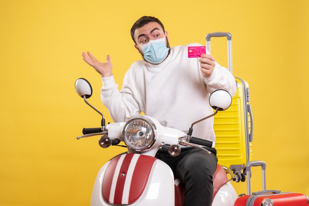 Vista frontal do conceito de viagem com um jovem preocupado numa máscara médica sentado na motocicleta com uma mala amarela e segurando o cartão do banco