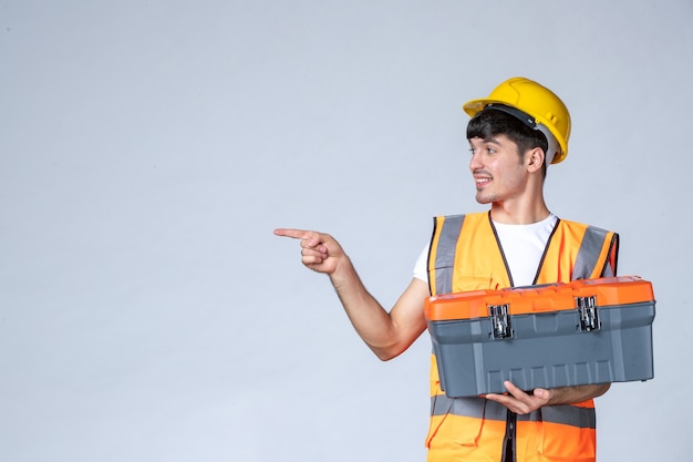 Vista frontal do construtor masculino de uniforme segurando uma caixa de ferramentas pesada na parede branca