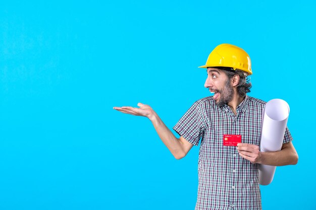 Vista frontal do construtor masculino segurando o plano e o cartão do banco sobre fundo azul construtor de projeto de dinheiro construção de trabalho capacete cor de trabalho