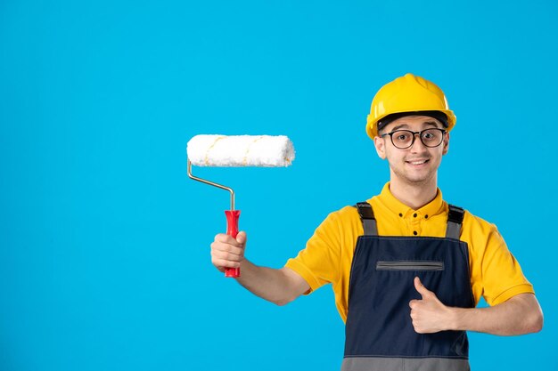 Vista frontal do construtor masculino sorridente de uniforme e capacete com rolo de pintura em azul