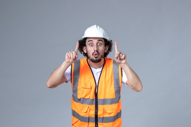 Vista frontal do construtor masculino surpreso usando colete de advertência com capacete de segurança e apontando para cima na parede de onda cinza