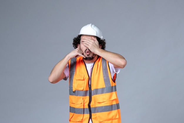 Vista frontal do construtor masculino usando colete de advertência com capacete de segurança e fazendo gesto de me ligar fechando os olhos na parede de onda cinza