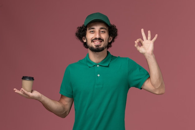 Foto grátis vista frontal do correio masculino com uniforme verde e capa segurando a xícara de café de entrega no fundo rosa uniforme serviço de entrega trabalho trabalhador trabalho masculino empresa