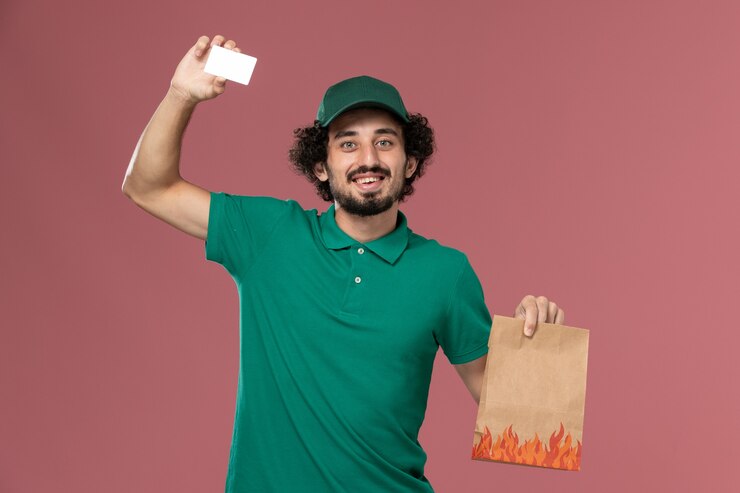 Foto grátis vista frontal do correio masculino com uniforme verde e capa segurando cartão e pacote de comida no fundo rosa serviço uniforme entrega trabalhador masculino trabalhador