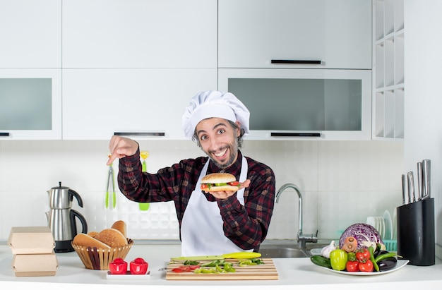 Vista frontal do cozinheiro masculino segurando hambúrguer apontando para o pão na mesa em pé atrás da mesa da cozinha