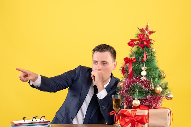 Vista frontal do dedo de um homem curioso apontando algo sentado à mesa perto da árvore de natal e presentes em amarelo