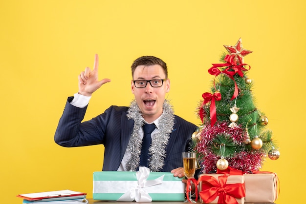 Vista frontal do dedo do homem feliz apontando para cima sentado à mesa perto da árvore de natal e presentes em amarelo