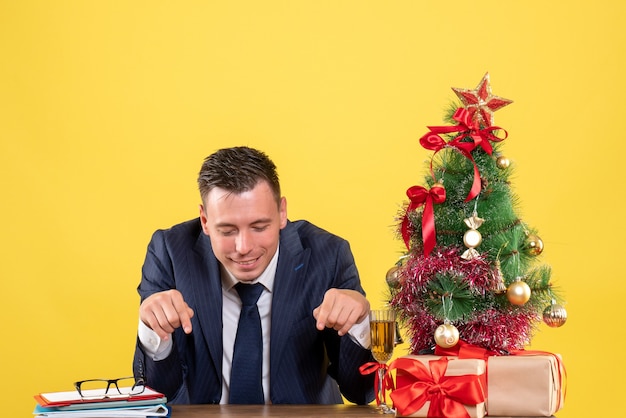 Vista frontal do dedo do homem sorridente apontando para a mesa sentada perto da árvore de natal e presentes em amarelo