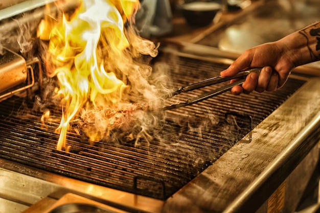 Vista frontal do delicioso bife suculento em chamas com fogo e fumaça na mão do chef profissional virando o conceito de bife de culinária e restaurante de comida e cozinha