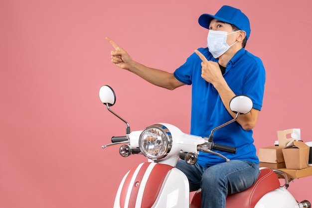 Vista frontal do entregador chocado com máscara médica e chapéu, sentado na scooter, apontando para cima no fundo cor de pêssego