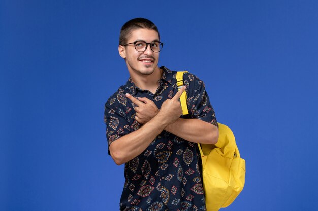 Vista frontal do estudante do sexo masculino com camisa de algodão escura e mochila amarela sorrindo na parede azul