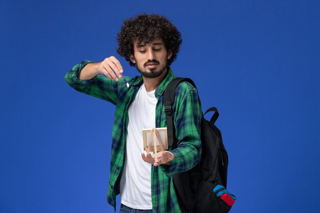 Vista frontal do estudante do sexo masculino com camisa quadriculada verde com mochila preta segurando borla e cavalete na parede azul