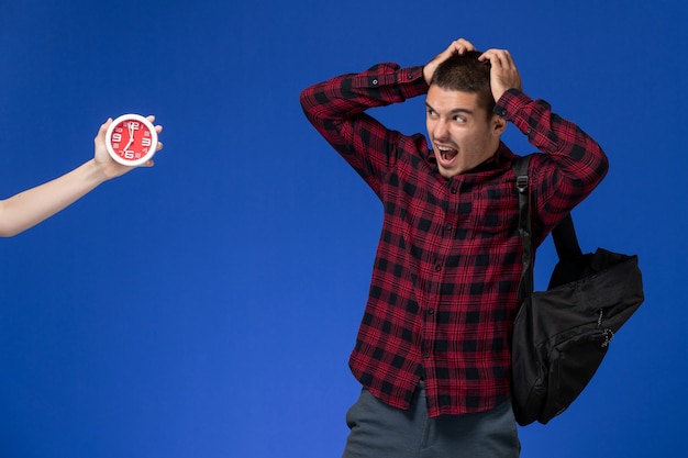 Foto grátis vista frontal do estudante do sexo masculino com camisa quadriculada vermelha com mochila preta com medo de relógios na parede azul