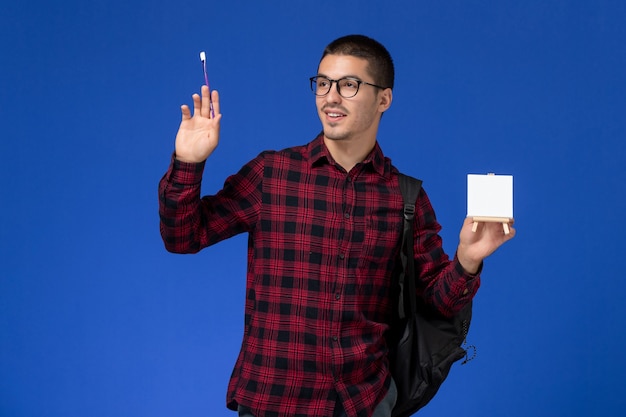 Vista frontal do estudante do sexo masculino com camisa quadriculada vermelha com mochila segurando a borla e cavalete na parede azul claro