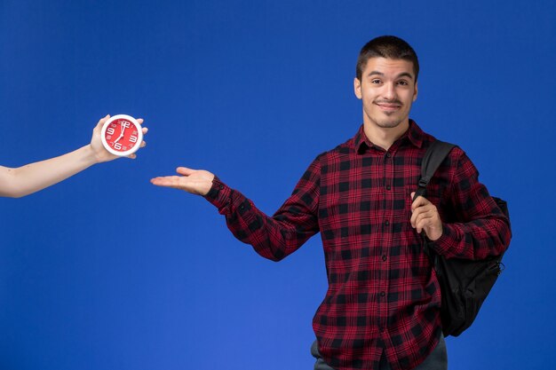 Vista frontal do estudante do sexo masculino com camisa quadriculada vermelha com mochila sorrindo na parede azul