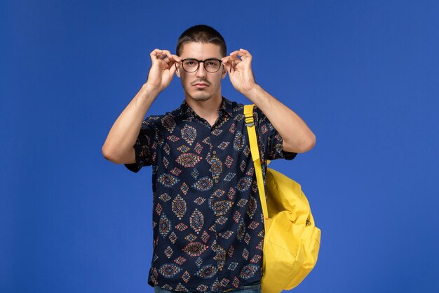 Vista frontal do estudante do sexo masculino com mochila amarela, posando e olhando para a câmera na parede azul