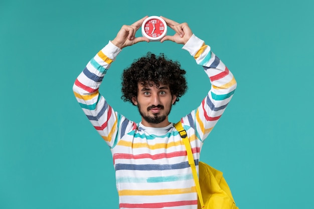 Foto grátis vista frontal do estudante do sexo masculino com mochila amarela segurando relógios na parede azul clara