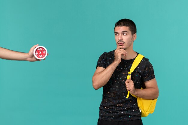Vista frontal do estudante do sexo masculino na mochila de camiseta amarela escura pensando na parede azul clara