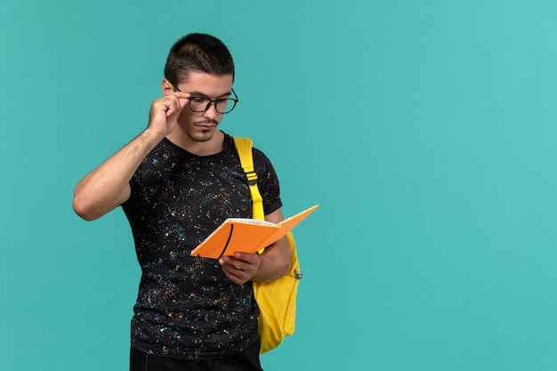 Vista frontal do estudante do sexo masculino na mochila de camiseta amarela escura segurando e lendo o caderno na parede azul