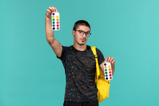 Vista frontal do estudante do sexo masculino na mochila de camiseta amarela escura segurando pinturas na parede azul